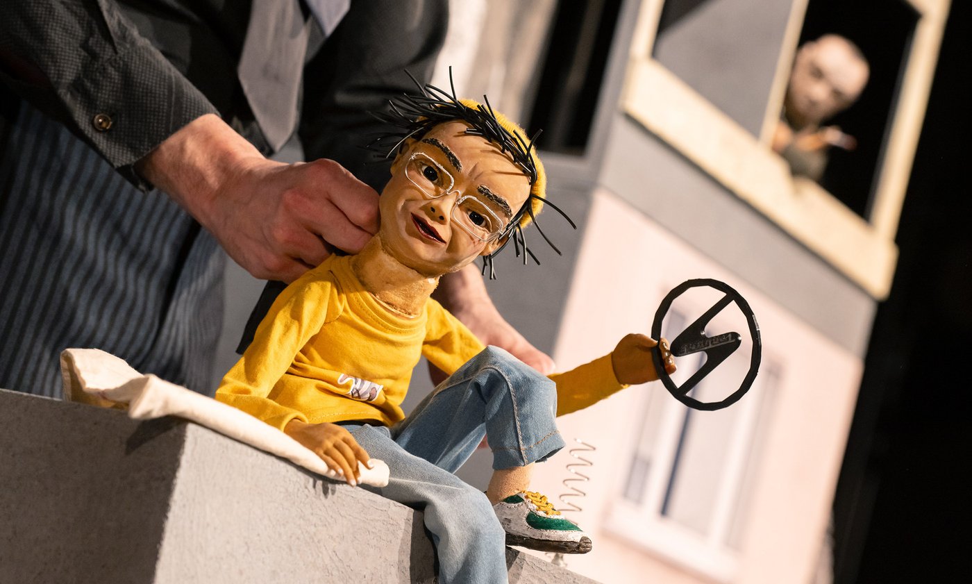 Theaterpuppe mit Trabant-Logo in der Hand, im Hintergrund ein Puppenspieler